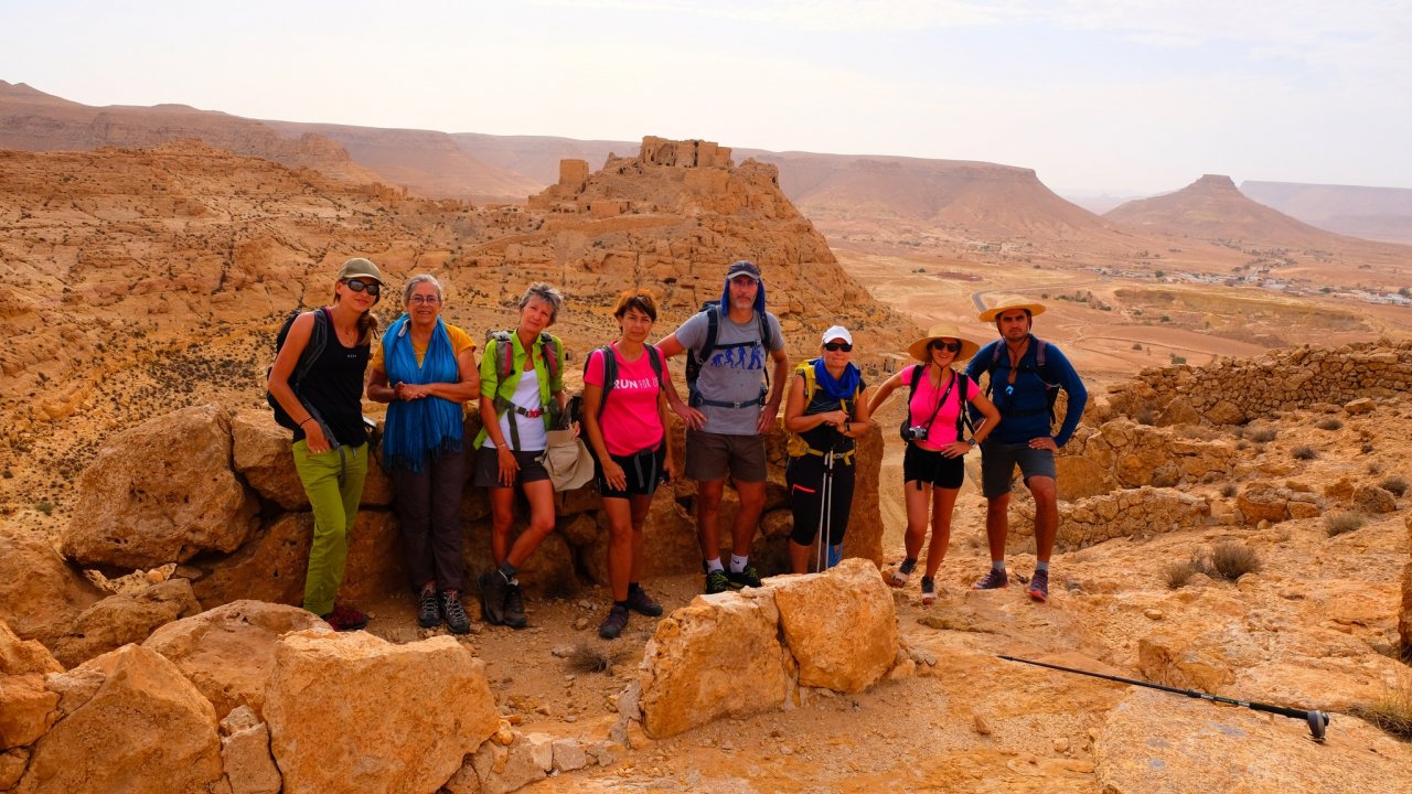 A group of trekkers, equipped with backpacks and hats, poses for a photo in front of the ancient Berber ksour ruins of Douiret, located in the rugged mountains of southern Tunisia