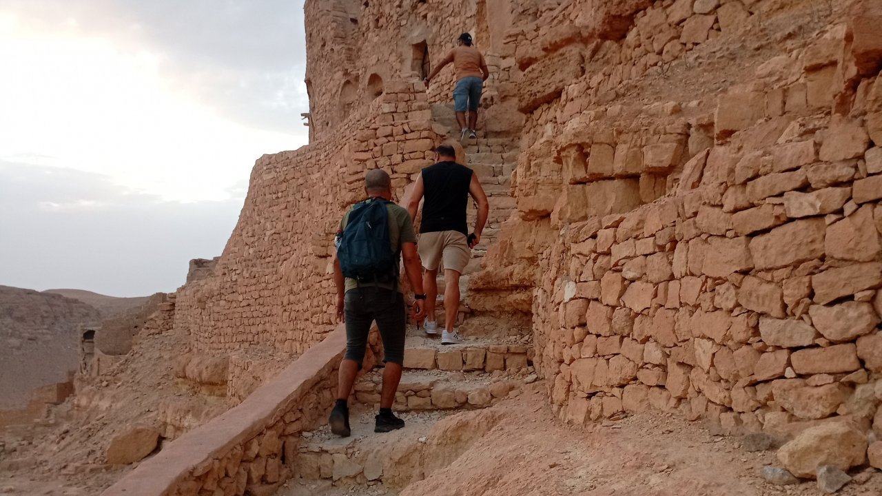 Three hikers ascend the winding, ancient stone steps carved into the cliff face, approaching the fortified Berber granary (ksar) at Douiret, Tunisia, as the sun sets.