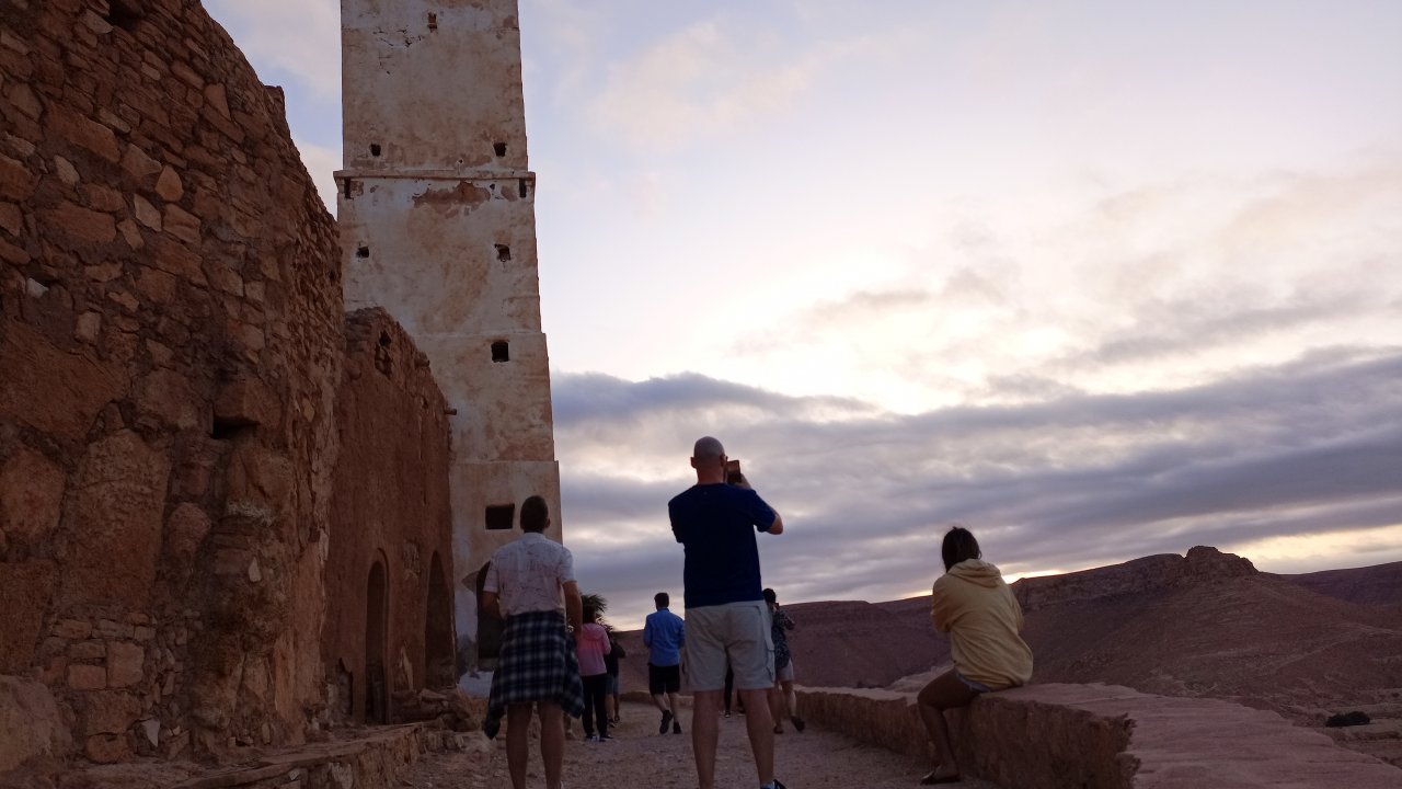 Tourists stand on a raised, ancient stone path near the fortified granary (ksar) of Douiret, Tunisia, taking in the panoramic mountain view at sunset.