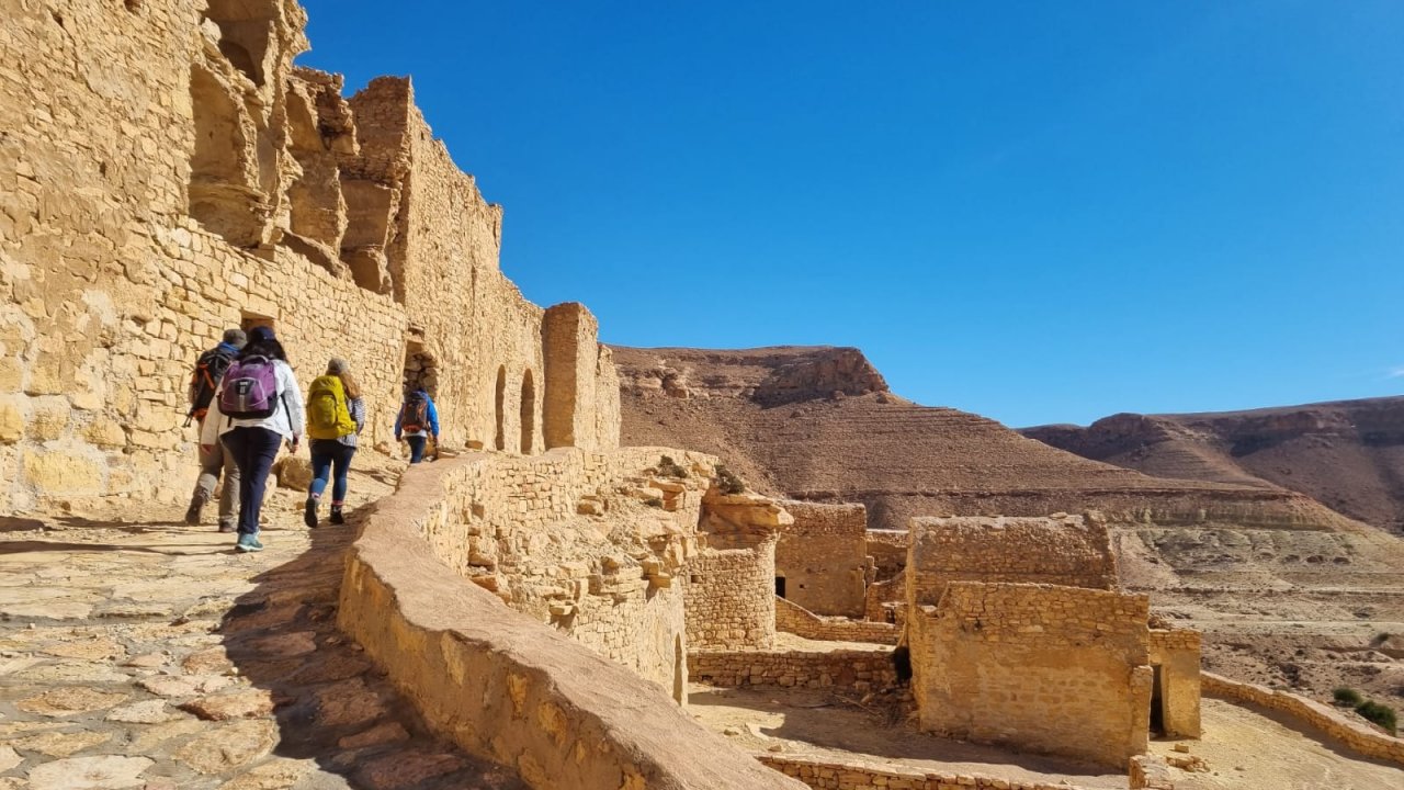 A group of trekkers, led by a guide, walks along a winding, elevated stone path, exploring the ancient, fortified Berber village ruins of Douiret in the Dahar region of southern Tunisia, under a clear blue sky.